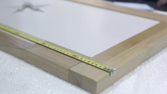 Close-up of worker measures wooden frame. Action. Control measurement of wooden frame. Worker of furniture factory checks dimensions of wooden frame with measuring tape