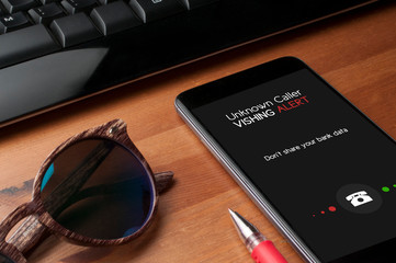 Vishing (voice phishing) concept, a smartphone on a table next to a computer keyboard and sunglasses show an unknower caller call with vishing alert and a reminder to not share personal bank data