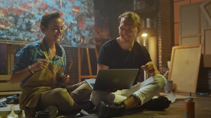 Couple of Artists Sitting in their Studio Use Laptop Computer, Working on a Project, Talking and Smiling. Two Painters Doing Research on Internet High Five after Thinking of Successful Idea. Workshop 