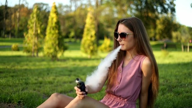 Vape teenager. Young pretty sporty caucasian girl in a pink jumpsuit and sunglasses smoking an electronic cigarette in the park on a sunny day in summer. Bad habit. Vaping activity.