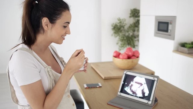 Beautiful young pregnant woman eating an ice cream while looking ultrasound of her baby in the digital tablet at home.