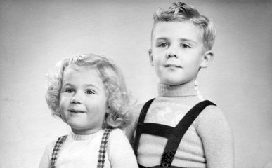 Early 1950s duo portrait of a young boy and girl with blond hair and curls.