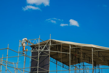 preparing for some event concept picture of stage installation by working person, pipes frame on blue sky background with empty space for copy or text