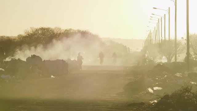 Late afternoon sunlight illuminates a road covered in smoke next to heaps of garbage smoldering in a large dump as people walk by, silhouettes visible in the distance.