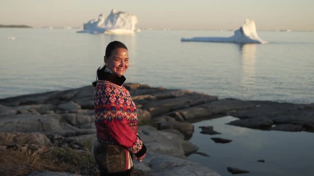 Portrait of smiling woman in traditional outfit standing on rock by Disko Bay during sunset