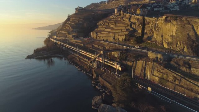 Train Goes Along Lake Geneva Shore at Sunset. Terraced Vineyards and Houses. Switzerland. Aerial View. Drone Follows Train