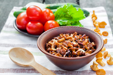 Fried chanterelles with onion in rustic bowl and plate with fresh vegetables for salad on background