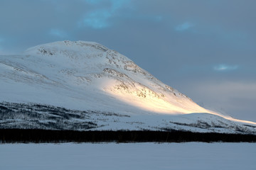 Snow covered mountain Gappetjahkka in Lapland Sweden with sunbeam on snowfield, cold grey sky