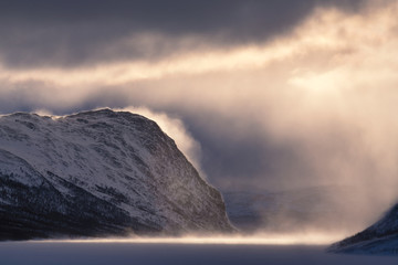 Dramatic nordic winter landscape in Lapland Sweden during snow storm with mystic light, sun, mist, and dark cloud layer