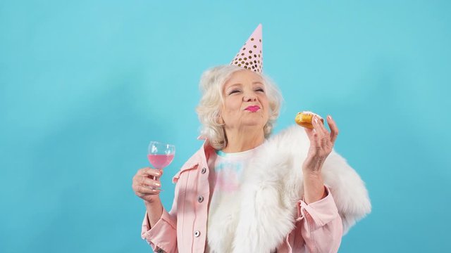 Glamour old lady in party hat holding a glass of wine and eating yummy cake. Isolated blue background. People, happiness