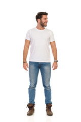 Handsome Man In Jeans And White T-shirt Is Standing, Looking Away And Smiling. Front View.