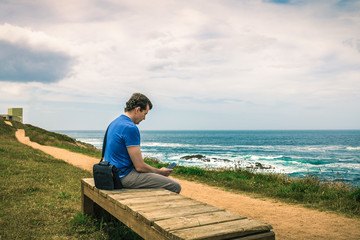 man sitting on a bench using smart phone facing the sea next to the pathway