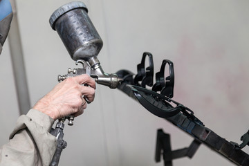 A male worker in jumpsuit and mask paints with a spray gun a part of the car interior element in...
