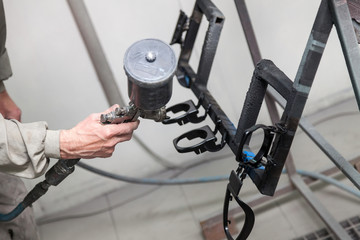 A male worker in jumpsuit and mask paints with a spray gun a part of the car interior element in black after being damaged. Auto service industry professions.
