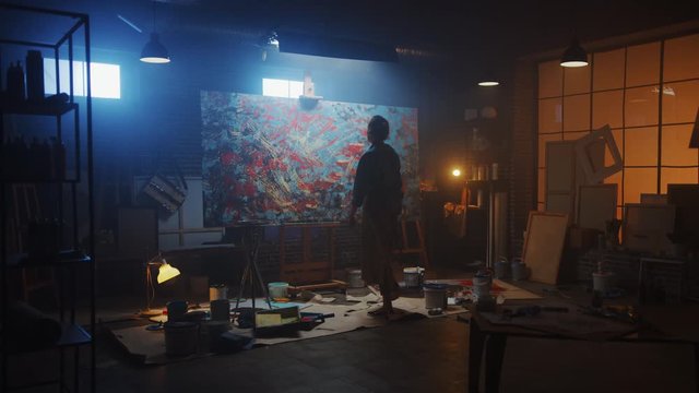 Talented Female Artist Works on Abstract Oil Painting, Using Paint Brush She Creates Modern Masterpiece. Dark and Messy Creative Studio where Large Canvas Stands on Easel Illuminated. Zoom out