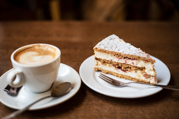 Delicious slice of cake with cottage cheese and cherry jam next to a cup of cappuccino on a wooden table