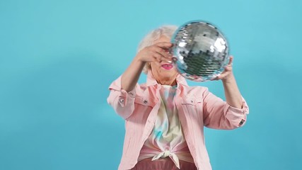 Funny old woman in sunglasses holding a disco ball, isolated yellow background. Lifestyle, woman wants to dance.
