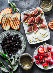 Mediterranean style snack appetizers - dried olives, figs, cheese, grilled bread, strawberries, raspberries and white wine on a dark background, top view