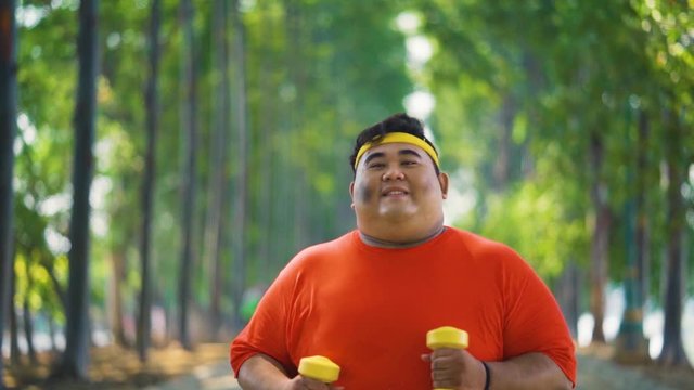 Slow motion of overweight young man jogging at the park while holding dumbbells. Shot outdoors