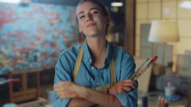 Beautiful Female Artist Dirty with Paint, Wearing Apron, Wipes Sweat from Forehead, Crosses Arms while Holding Brushes, Satisfied She Looks at Camera with a Smile. Authentic Creative Studio