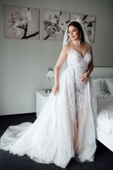 Portrait of beautiful bride in dress and long veil standing near bed in bedroom. Bride's morning preparation. Wedding concept. Beautiful young bride with wedding makeup and hairstyle in bedroom.