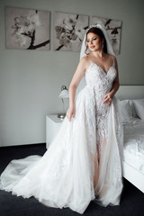 Fototapeta premium Portrait of beautiful bride in dress and long veil standing near bed in bedroom. Bride's morning preparation. Wedding concept. Beautiful young bride with wedding makeup and hairstyle in bedroom.