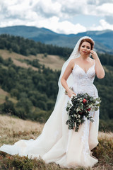 Fototapeta na wymiar Happy smiling bride in white stylish dress, with a wedding bouquet, beautiful mountains landscape in the background. Summer wedding, honeymoon trip countryside. Natural makeup, hairstyle