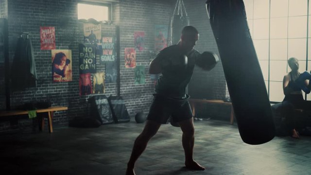 Strong and Handsome Male Kickboxer is Working Out in a Loft Gym with Motivational Posters on Walls. Masculine Athlete is Practising Punches on a Boxing Bag. He's Serious and Energetic.