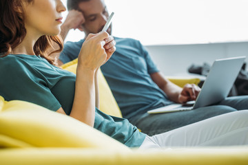 partial view of woman using smartphone while sitting near husband using laptop