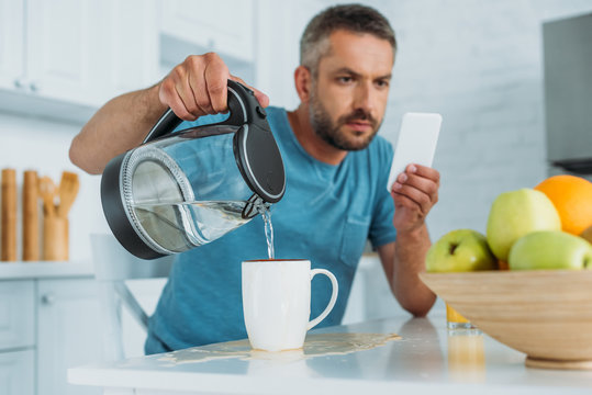 selective focus of man overfilling cup with water while sitting at kitchen table and using smartphone
