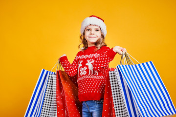 Excited adorable child in red Santa hat and sweater with deer carrying colorful shopping bags with Christmas gifts on yellow background