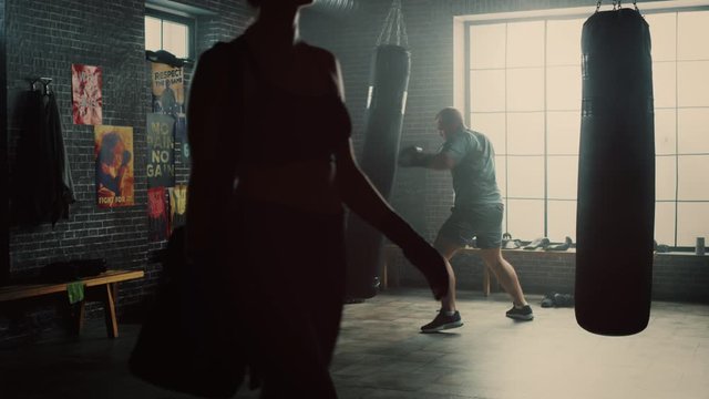 Strong and Handsome Male Kickboxer is Working Out in a Gym with Motivational Posters on Walls. Masculine Athlete is Practising Punches on a Boxing Bag. Fit Girl is Passing By.