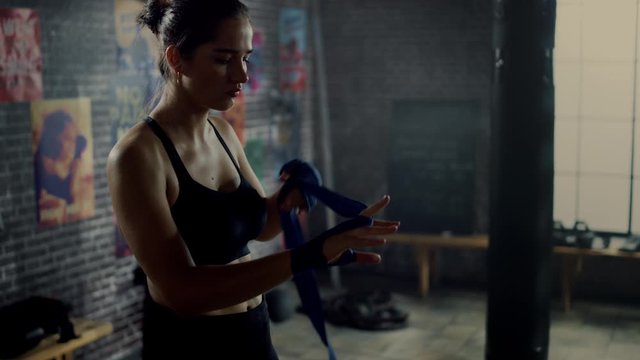 Strong and Beautiful Female Wrapping Her Hands with Handwraps to Start Her Kickboxing Workout in a Gym with Motivational Posters on Walls. She Uses Blue Straps Around Her Wrist, Thumb and Knuckles.
