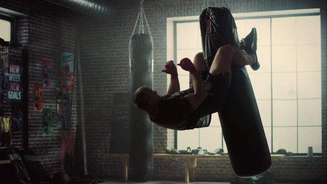 Insane Punching Bag Crunches. Strong Masculine Man Embrassing the Bag with His Legs and Pumping His Abs and Core Muscles. Handsome Male Trains in a Gym with Motivational Posters.