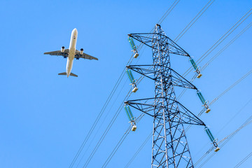 View from below of an airliner in landing approach flying over a high-voltage power line against blue sky with an electricity pylon in the foreground.