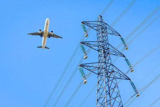 View from below of an airliner in landing approach flying over a high-voltage power line against blue sky with an electricity pylon in the foreground.
