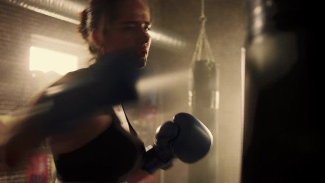 Fit Athletic Woman Kickboxer Punches and Hits the Punching Bag During a Workout in a Loft Gym with Motivational Posters. She's Exhausted and Tired After Her Intense Self-Defence Training. Warm Light.