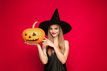 Young blonde woman in black hat and costume on red background. Attractive caucasian female model. Halloween, black friday, cyber monday, sales, autumn concept. Copyspace. Holds pumpkin, smiles.