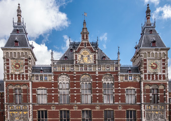 Fototapeta na wymiar Red brick with white trim upper part facade Centraal Railway Station with towers, clocks, and plenty of frescoes under blue sky with white clouds.