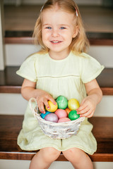 Fototapeta premium Cute little child girl with blonde hair on Easter day. Girl holding basket with painted eggs and sitting on the stairs at home