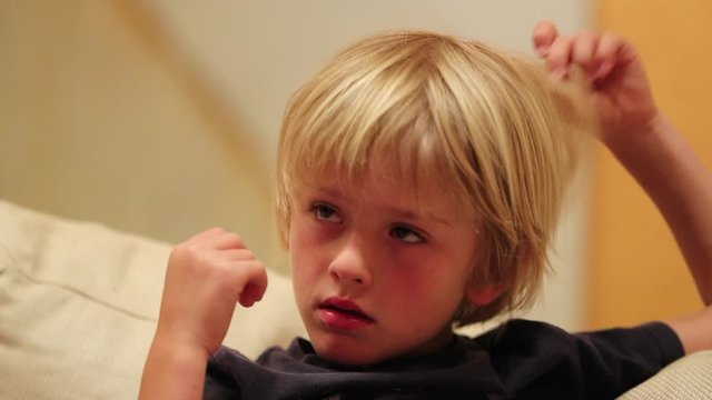Small boy playing with hair while watching TV screen hypnotized by content