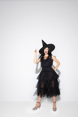 Young brunette woman in black hat and costume on white background. Attractive caucasian female model. Halloween, black friday, cyber monday, sales, autumn concept. Copyspace. Pointing, posing.