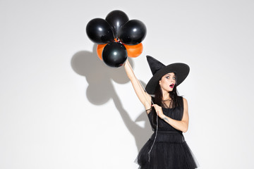 Young brunette woman in black hat and costume on white background. Attractive caucasian female model. Halloween, black friday, cyber monday, sales, autumn concept. Copyspace. Holds balloons, shocked.