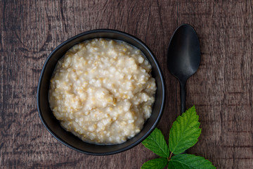 Hot cooked oatmeal in a black ceramic bowl on a wood table, with a black metal spoon and blackberry leaves next to bowl