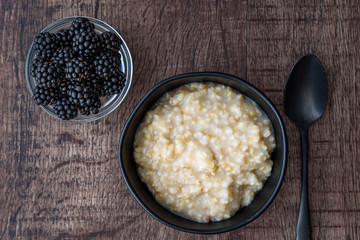 Hot cooked oatmeal in a black ceramic bowl on a wood table, with a small glass bowl of fresh blackberries and a black metal spoon next to bowl
