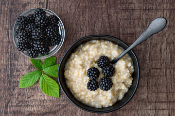 Hot cooked oatmeal with blackberries on top in a black ceramic bowl on a wood table, with a black metal spoon in the bowl and a small glass bowl of fresh blackberries next to bowl