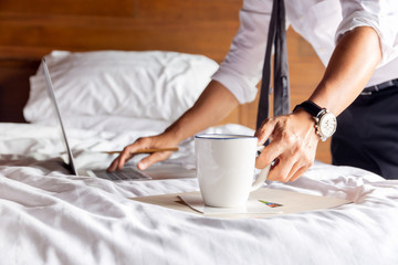 Businessman hand holding mug of coffee and working on laptop in bedroom.
