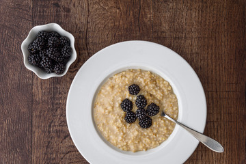 Cooked oatmeal in a white bowl with blackberries on top, small side bowl of fresh blackberries, on a wood table
