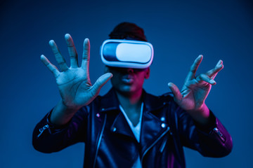 Portrait of young african-american woman's playing in VR-glasses in neon light on blue background. Concept of human emotions, facial expression, modern gadgets and technologies. Touches something in