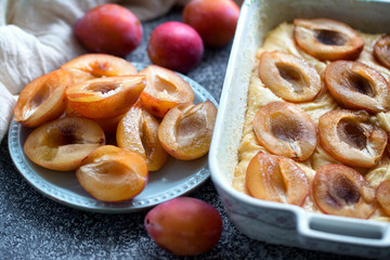 Dough with halves of fresh plums in ceramic shape, and cut plums on a blue plate on a grey table. Preparation for baking Plum Torte. 